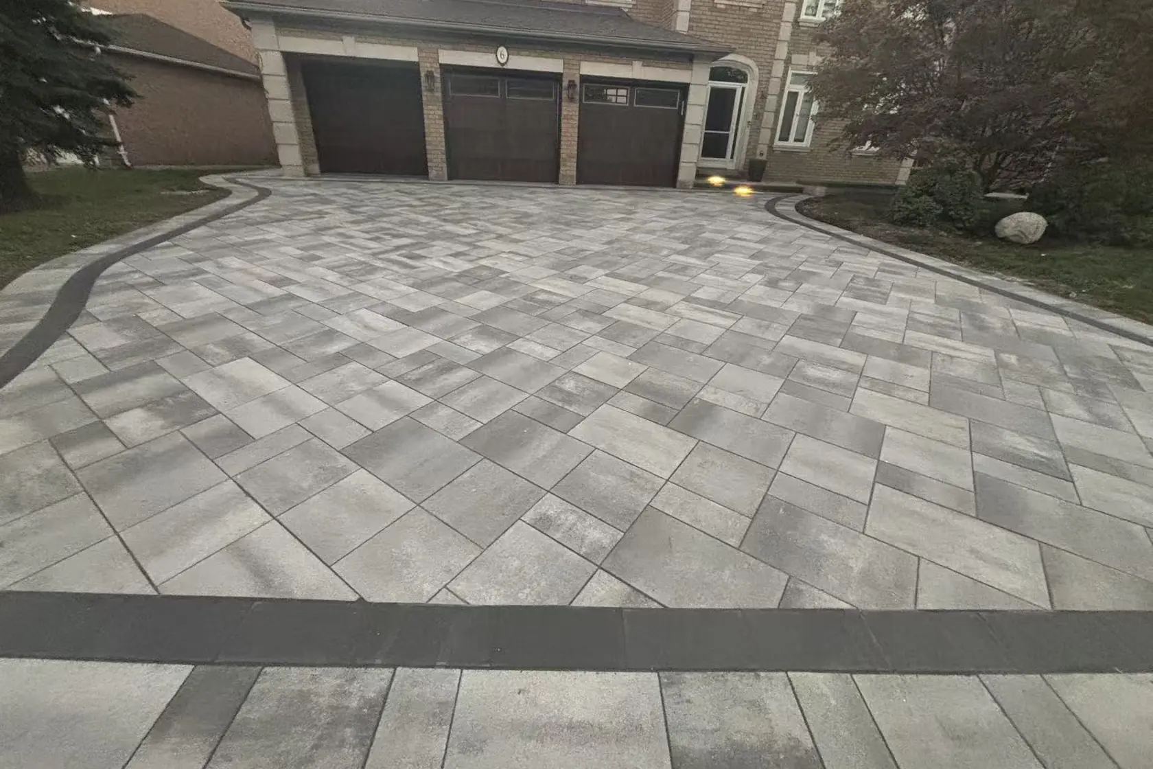 Grey interlock driveway -- large-format pavers, dark charcoal curved border, GTA brick home with double garage